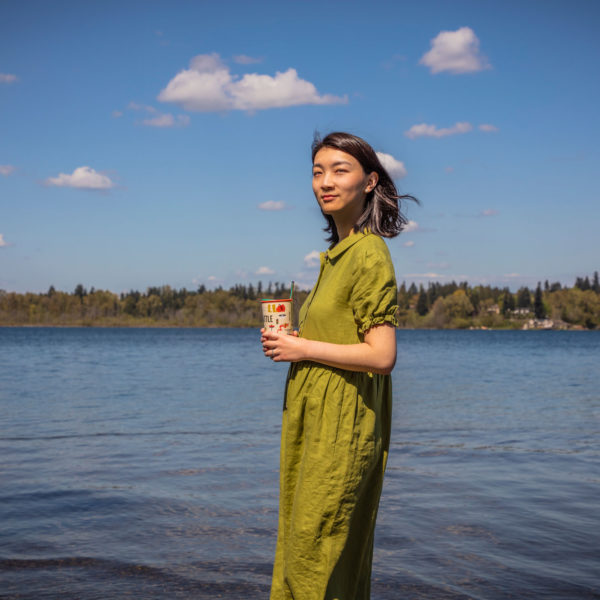 A woman in a green dress who is holding a coffee cup and standing at the shore of a lake