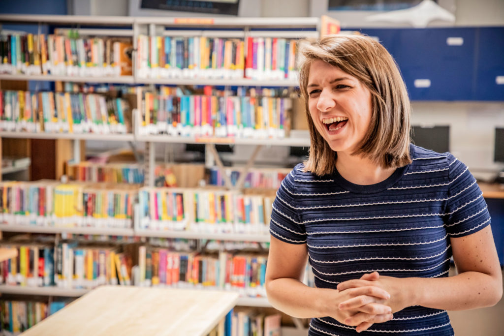 A woman smiling while standing in a school library with a shelf of books behind her
