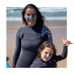 A woman and a little girl stand on the beach in wet suits making the sign for 