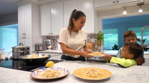 A woman stands in her kitchen and prepares plates of pasta.