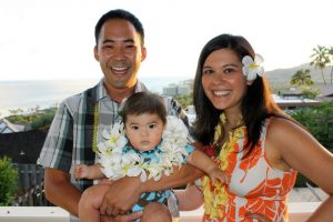 A man, woman, and child stand on a balcony in Hawaii with the ocean seen behind them