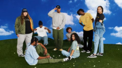 Group of eight people wearing Hardwear clothing, sitting and standing on grass, in front of a blue and white background.
