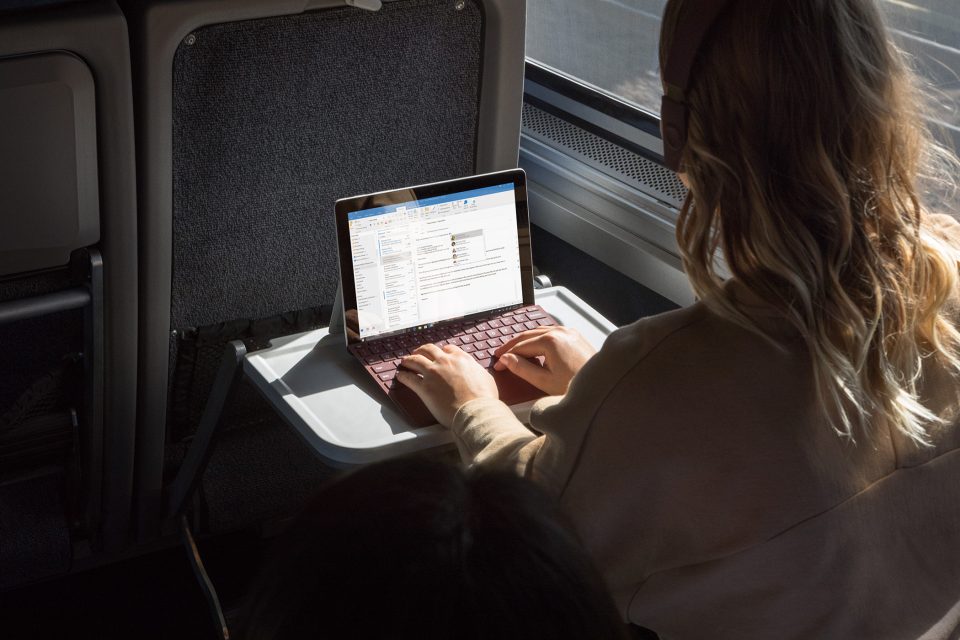 Junge Frauen im Zug arbeitet mit dem Surface Go an Outlook E-Mail, während sie Kopfhörer trägt.