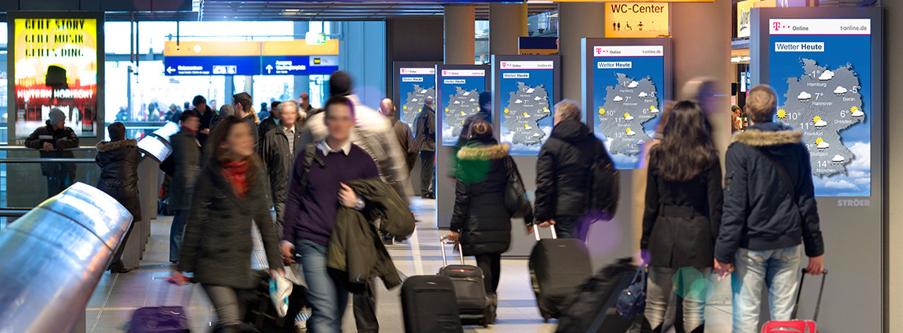 Passanten in einem Bahnhof laufen an bewegtbild Werbeflächen vorbei