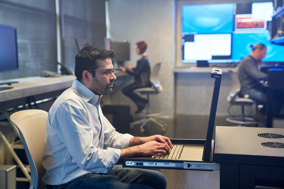 Mann mit Kragenhemd, der auf einer Serverstation in einem Sicherheitsraum arbeitet. Beschäftigte und große Monitore sind im Hintergrund zu sehen.