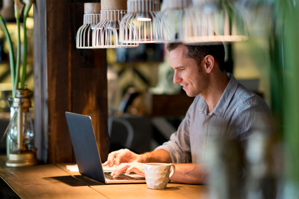 Ein Mann arbeitet an einem Laptop, er sitzt an einem Holztisch, über ihm hängen Lampen.
