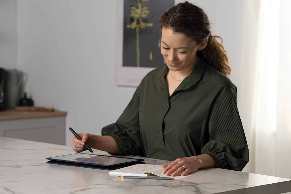 Eine Frau steht an einem Tisch und trägt Informationen von einem Notizbuch in ein Tablet um.