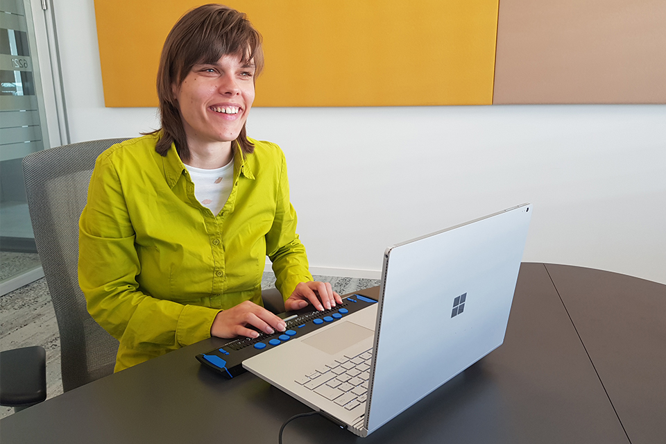 Eine Frau sitz vor einem Laptop mit Braillezeile, der auf einem Tisch steht