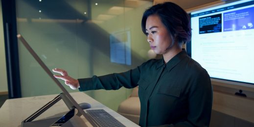 Eine Frau blickt konzentriert auf einen großen Bildschirm, den sie mit ihren Fingern berührt. Im Hintergrund ist ein großer Monitor mit Daten zu sehen.
