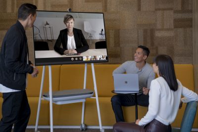 Drei Personen befinden sich in einem Raum, sie blicken auf auf ein Surface Hub, auf dem eine Teams-Besprechung stattfindet.