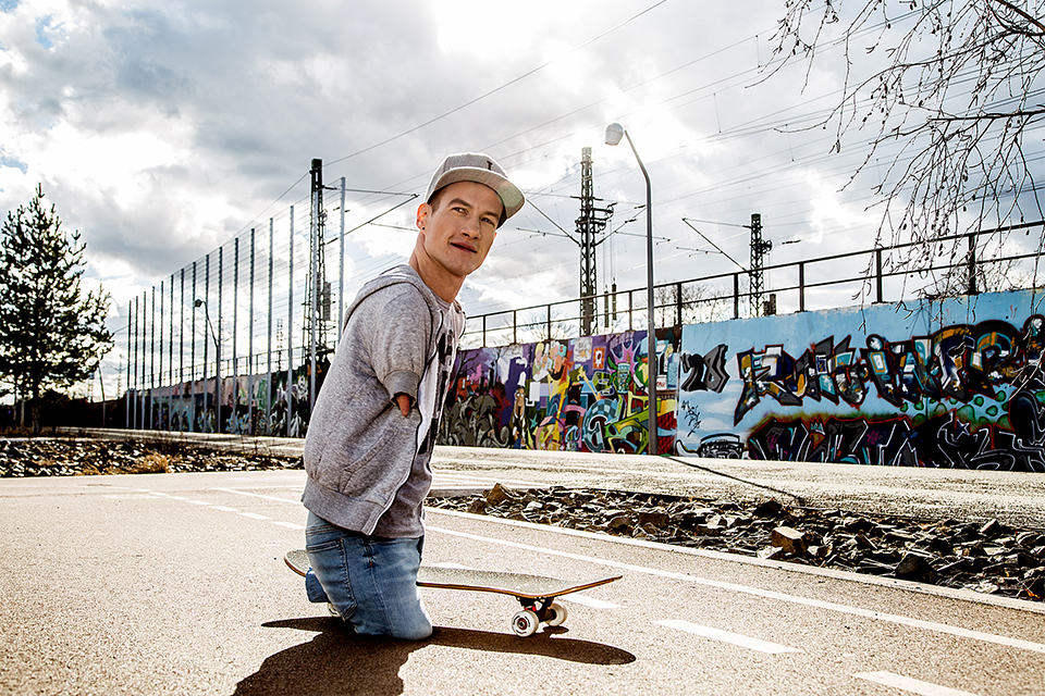 Ein Mann mit Behinderung fährt auf einem Skateboard. Im Hintergrund eine Mauer mit Graffiti