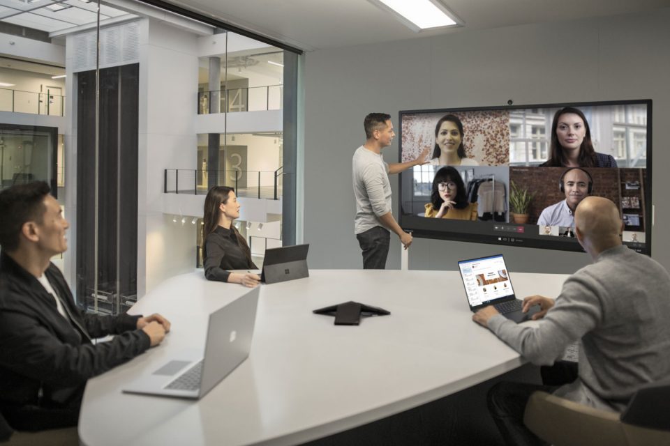 Ein Konferenzraum, in dem vier Personen mit ihren Laptops sitzen. An der Wand ist ein Surface Hub montiert, der eine Teams-Besprechung überträgt.