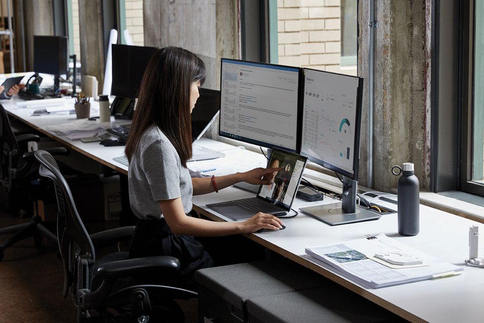 Eine Frau sitzt an einem Schreibtisch im Büro, vor ihr stehen zwei Monitore sowie ein Laptop, mit dem sie arbeitet.