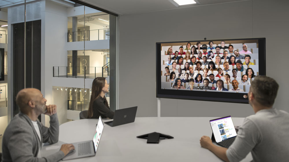 Ein Konferenzraum mit mehreren Personen, die auf Abstand sitzen. Auf dem Surface Hub an der Wand läuft eine Teams-Besprechung mit vielen Teilnehmenden. Sie nutzen den Zusammen-Modus.