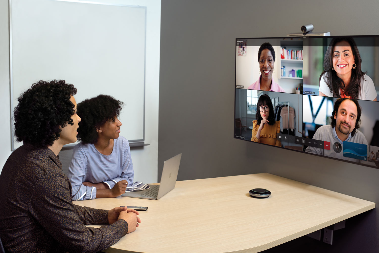 Zwei Menschen sitzen in einem kleinen Konferenzraum, sie blicken auf einen Bildschirm an der Wand, auf dem vier Teilnehmer*innen der Teams-Besprechung zu sehen sind.