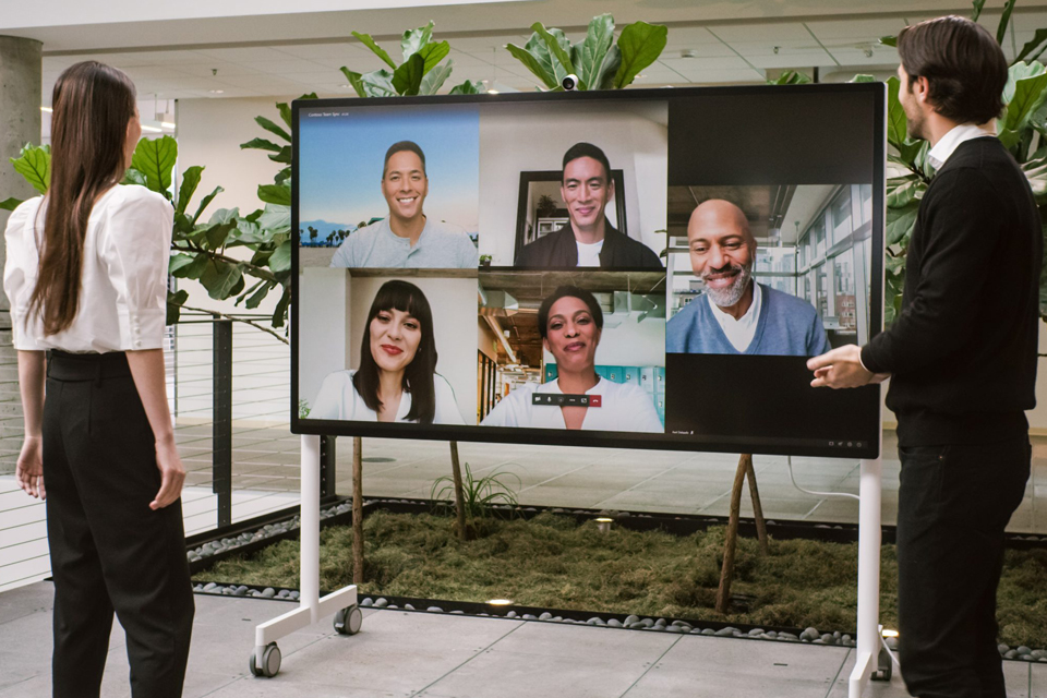 Eine Frau und ein Mann stehen vor einem Surface Hub 2S und nehmen an einem hybriden Teams-Meeting teil