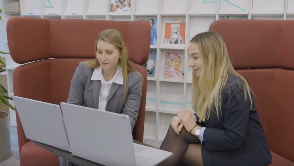 Zwei Frauen sitzen an ihren Laptops und unterhalten sich.