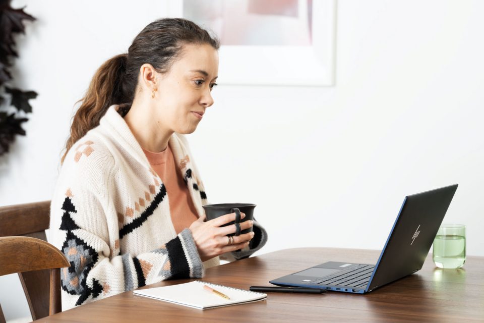 Eine Frau sitzt an einem Tisch, sie hält mit beiden Händen eine Tasse in der Hand. Vor ihr steht ein Laptop, auf den sie blickt.