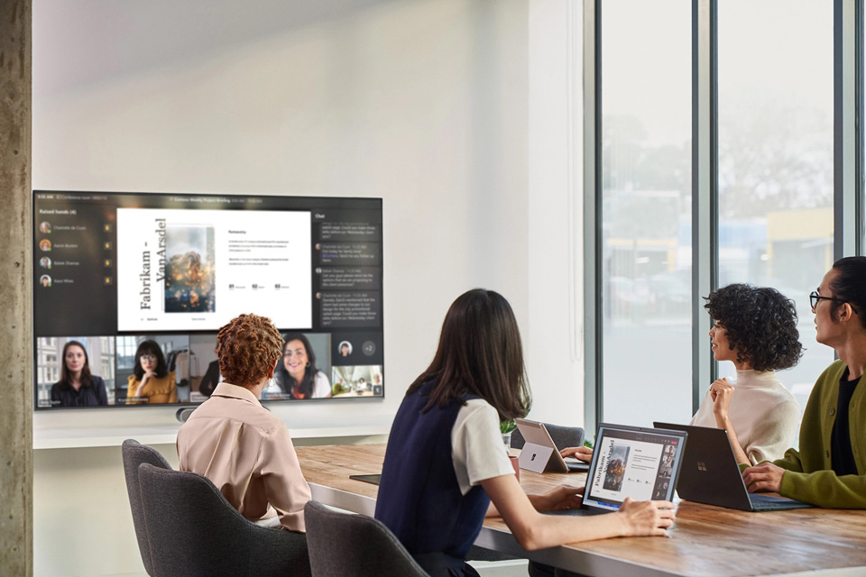 Personen sitzen in einem Konferenzraum und schauen auf einen Monitor an der Wand. Darauf zu sehen ist eine Teams-Besprechung, in der die virtuellen Teilnehmer*innen auf Augenhöhe gezeigt werden.