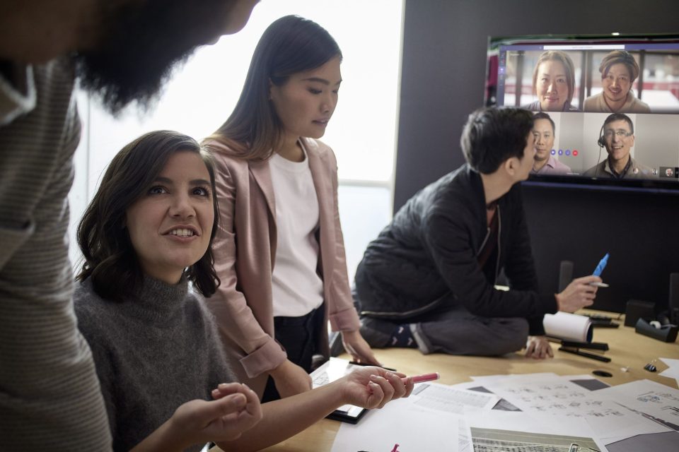 Eine Arbeitsgruppe aus drei Frauen hält ein hybrides Meeting ab. Auf dem Bildschirm im Hintergrund sind die Kolleg*innen zugeschaltet.