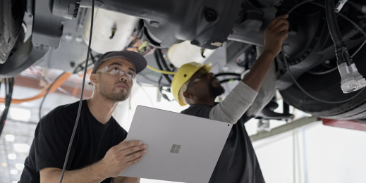 Zwei Männer arbeiten unter einem aufgebockten Auto. Sie tragen Schutzhelme und -brillen. Einer von ihnen hält ein Surface Book 2 in der Hand, während der andere montiert.
