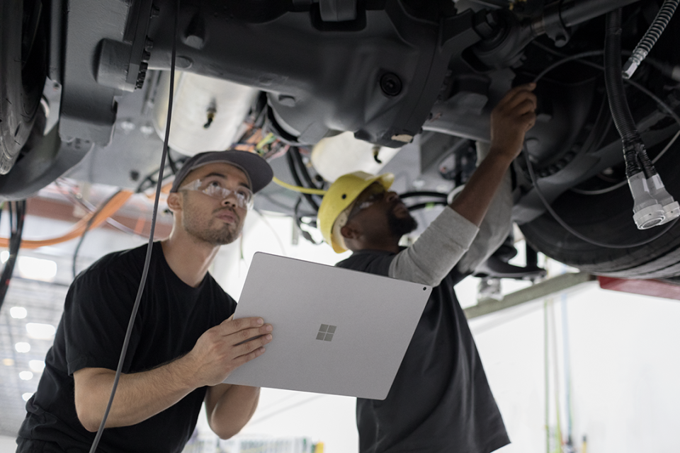 Zwei Männer arbeiten unter einem aufgebockten Auto. Sie tragen Schutzhelme und -brillen. Einer von ihnen hält ein Surface Book 2 in der Hand, während der andere montiert.