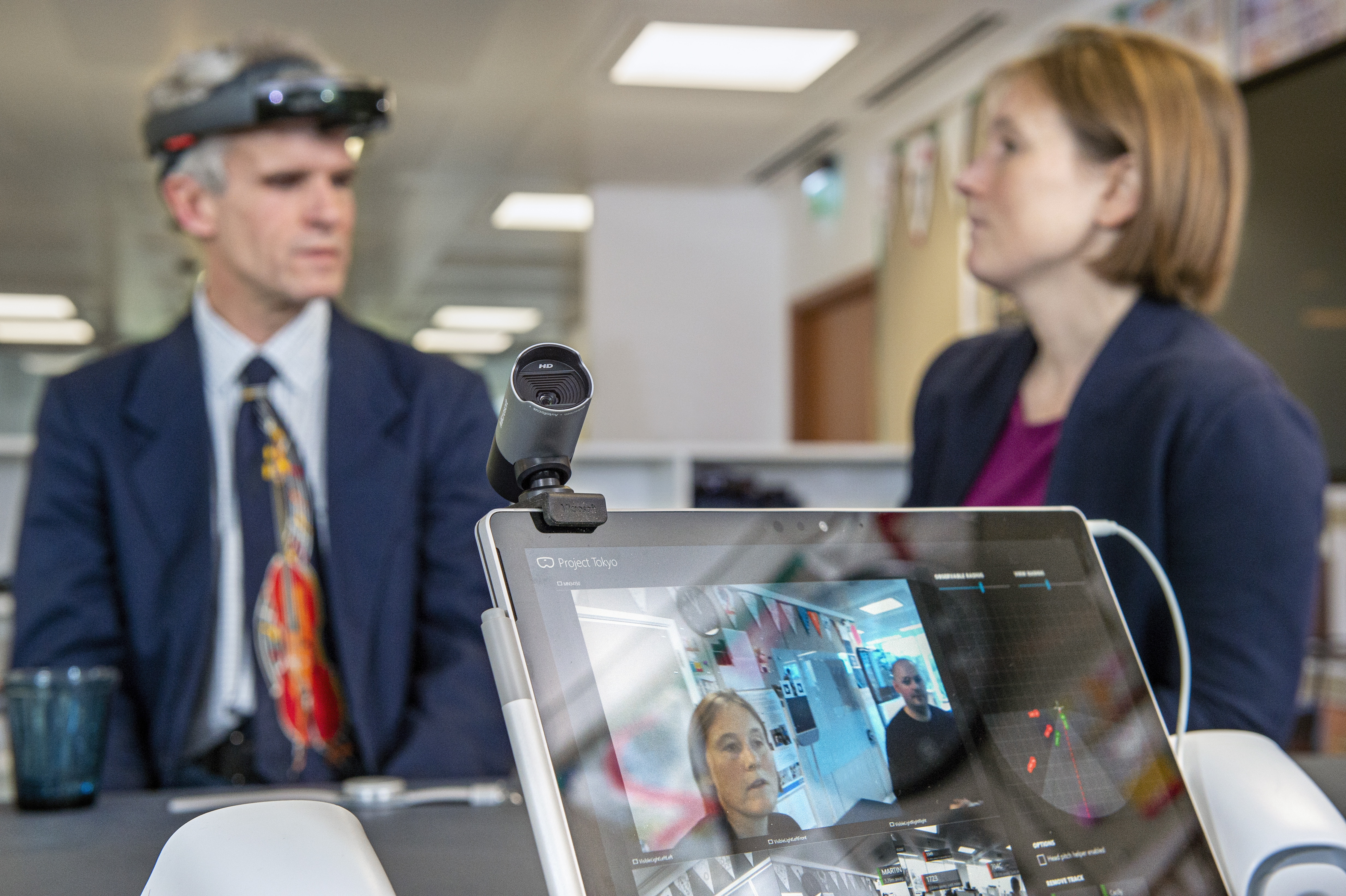 Una pantalla de computadora en primer plano muestra un tablero de Project Tokyo que representa el campo visual del dispositivo HoloLens, utilizado por Peter Bosher al fondo