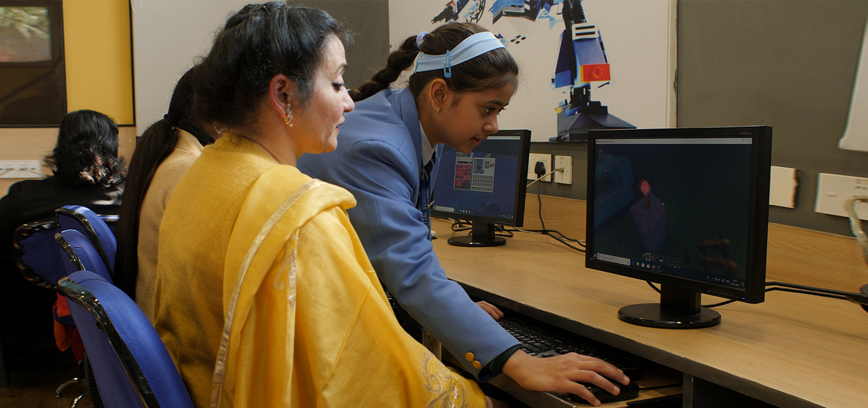 Una estudiante de 13 años con uniforme escolar ayuda a su profesora con Minecraft. Sus dos manos trabajan en el teclado mientras la profesora mira el monitor en la mesa. Hay dos otras mujeres y un monitor más en la imagen.