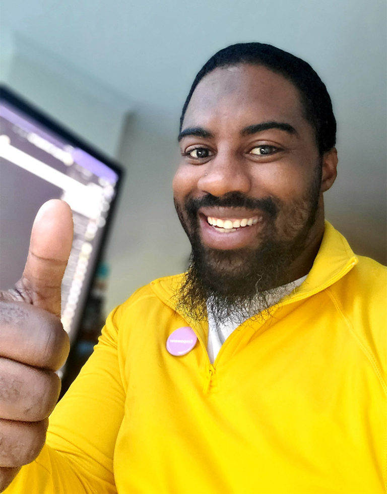Ethaniel Sinclair utiliza una playera amarilla de Microsoft Store mientras sonríe, levanta los pulgares y trabaja desde casa