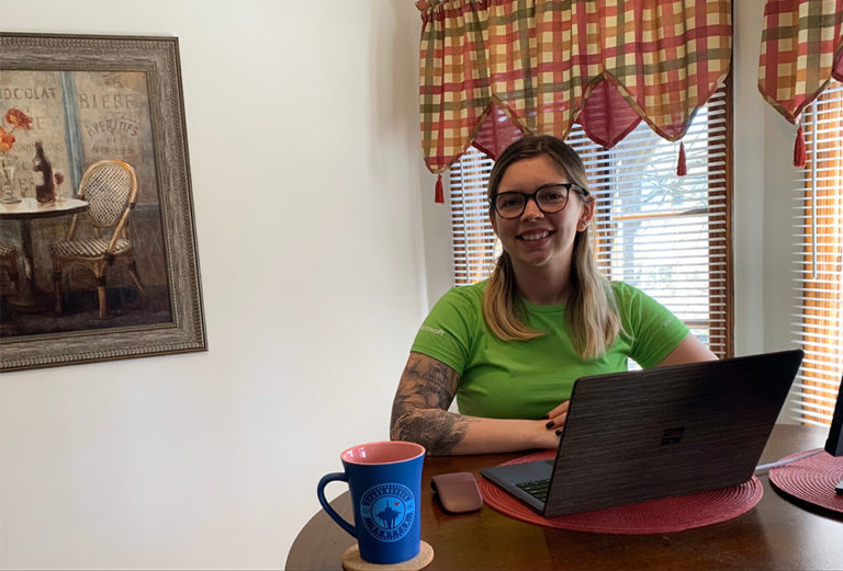 Samantha Janiec usa una playera verde de Microsoft Store y sonríe frente a su laptop mientras trabaja desde casa