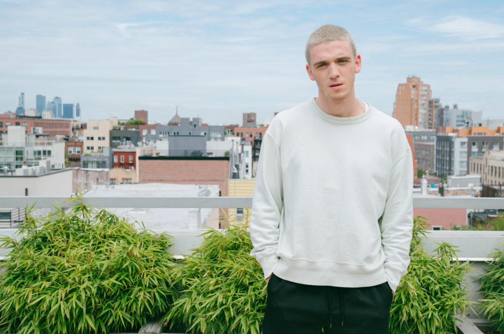 Lauv en un techo en Williamsburg, Brooklyn, mira a la cámara. De fondo, se aprecia Brooklyn. Lauv usa una sudadera blanca y pantalones deportivos negros.