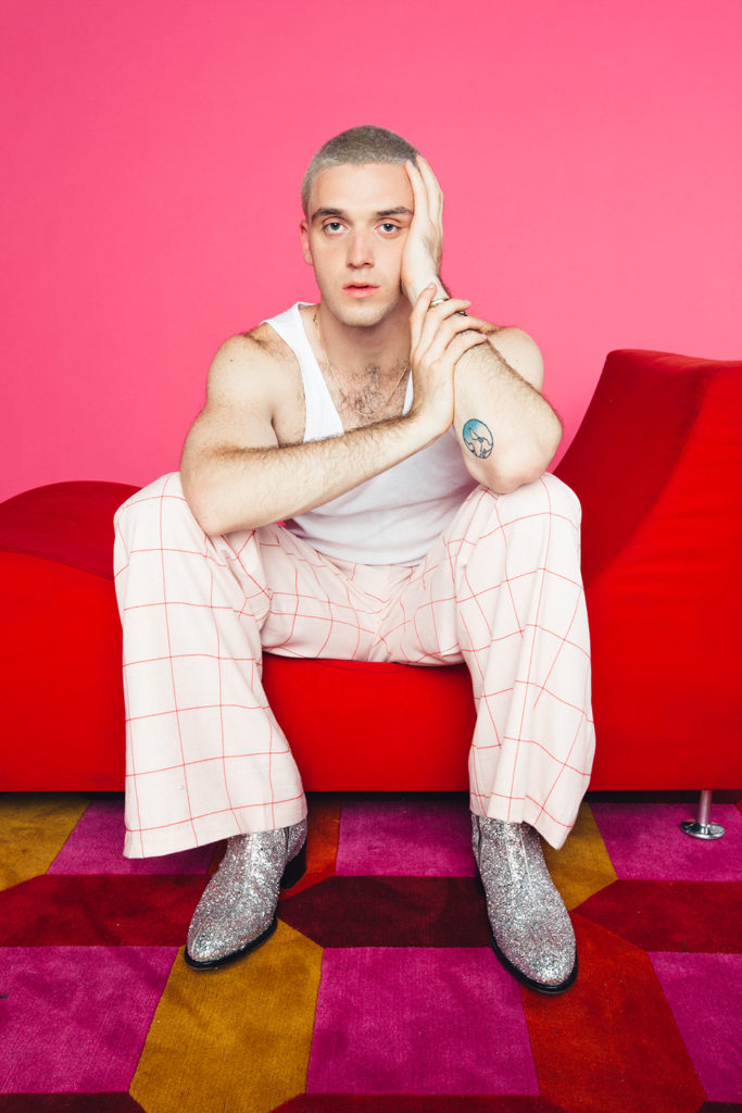 Lauv sentado en un sofá rojo con su codo izquierdo sobre su rodilla y su cabeza en su mano izquierda. Usa una playera blanca con pantalones a cuadros rosas y botas Chelsea con brillos plateados.