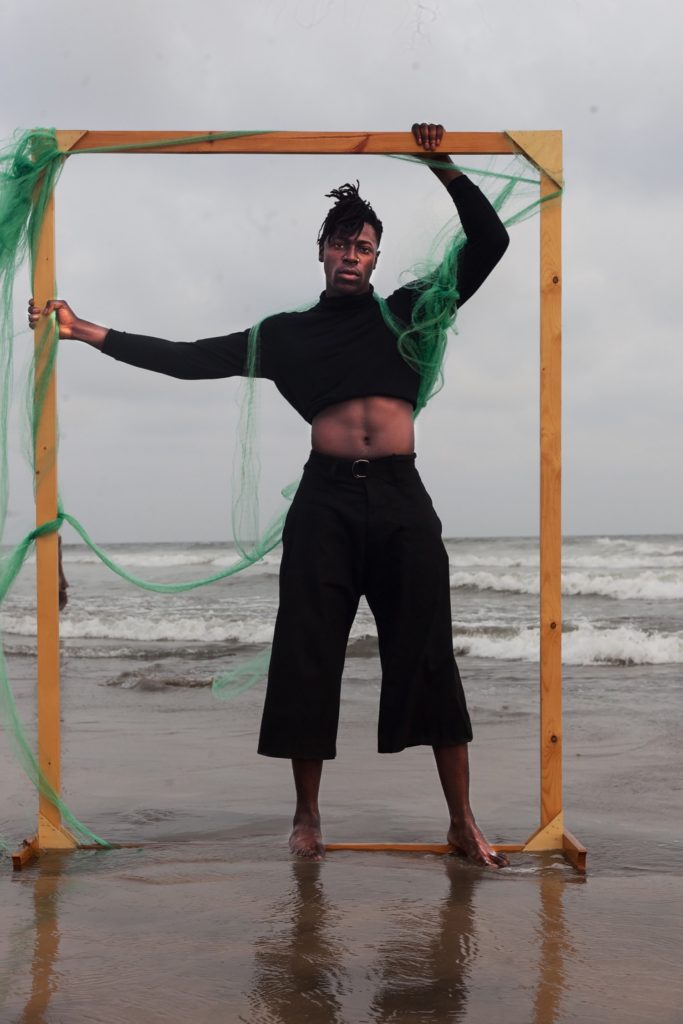 El músico Moses Sumney de pie, descalzo en una playa dentro de un marco alto rectangular de madera, con tul verde brillante unido. Usa un suéter de cuello de tortuga alto recortado negro y pantalones negros recortados de pierna ancha.