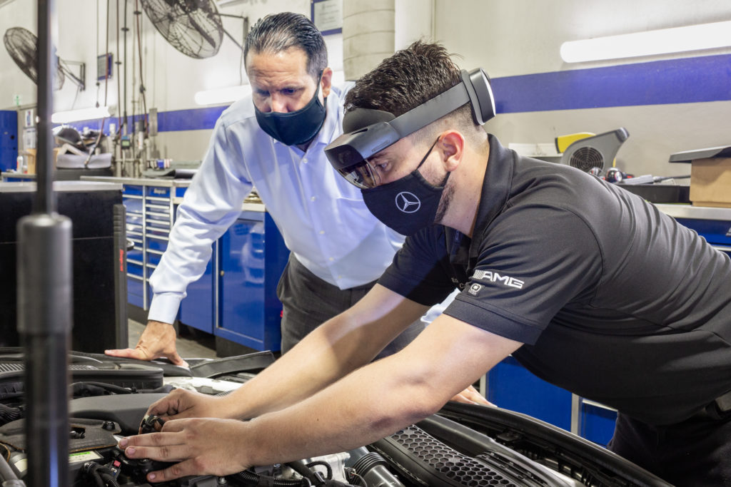 Un técnico en reparación automotriz que utiliza HoloLens 2 y un cubrebocas trabaja con sus manos en el motor de un Mercedes-Benz, mientras su supervisor, también con cubrebocas, observa por encima de su hombro.