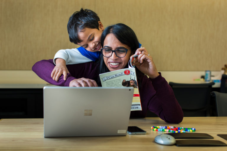 Una mujer sonríe y trata de trabajar en su laptop mientras su hijo juega detrás de ella