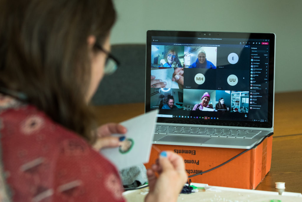 Una mujer añade cuentas a un trabajo artesanal mientras mira la pantalla de una laptop que muestra a gente que hace el mismo trabajo en Microsoft Teams.