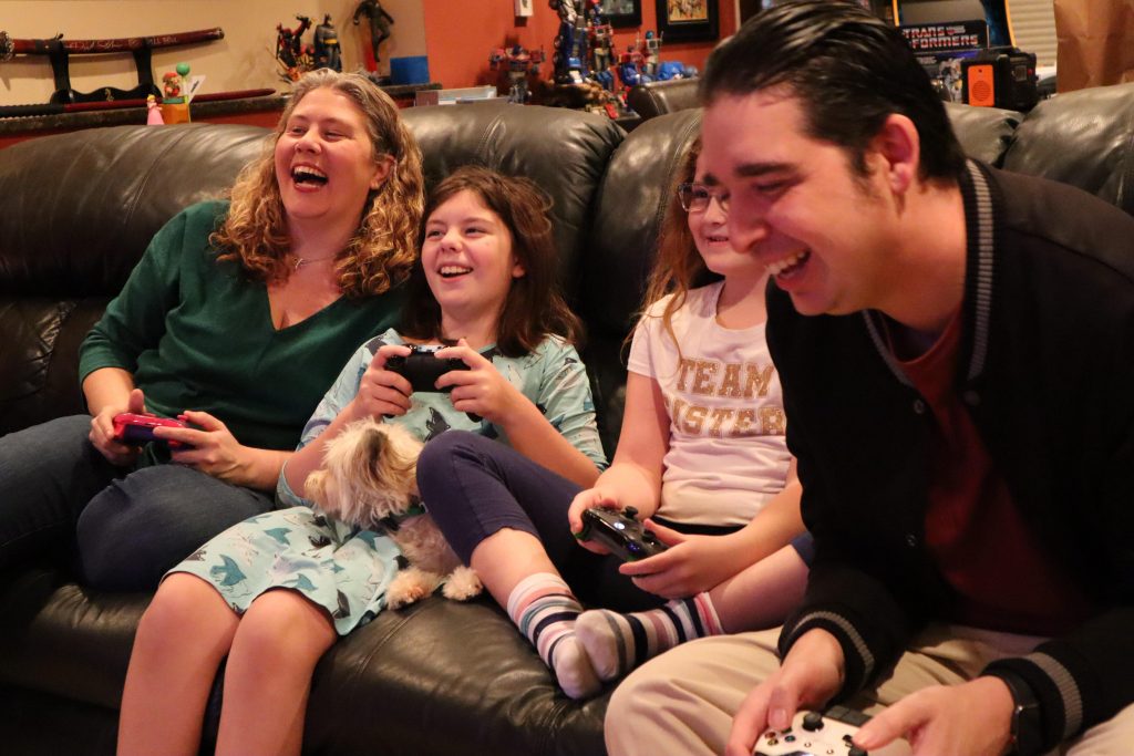 Una mujer, sus dos hijas y su esposo ríen en un sofá mientras juegan videojuegos
