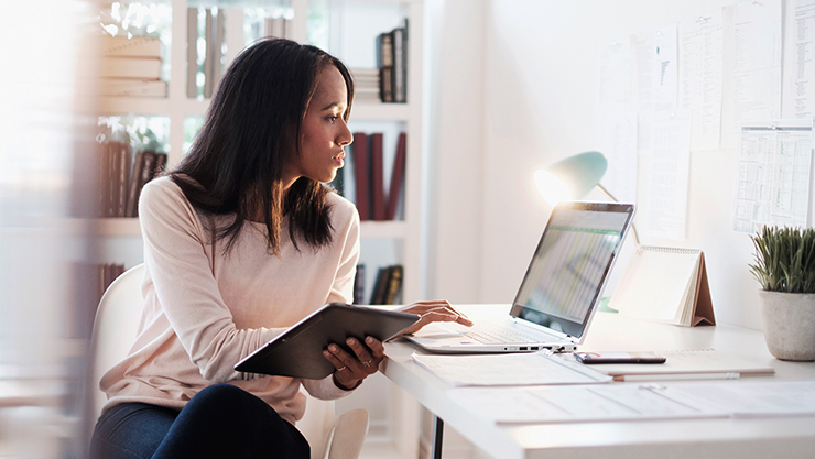 Mujer sostiene una libreta abierta mientras trabaja en una laptop en su oficina en casa