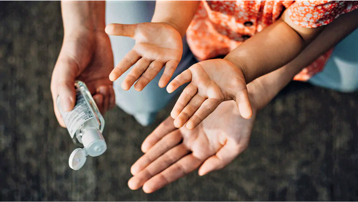 Se muestran dos manos de niños boca arriba y una mano de adulto boca arriba mientras otra mano de adulto sostiene una botella de desinfectante de manos.