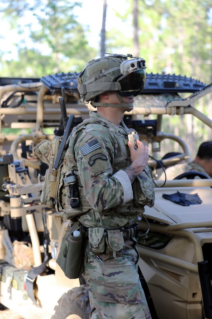 Foto de un soldado parado frente a un vehículo militar y con una diadema de realidad mixta.
