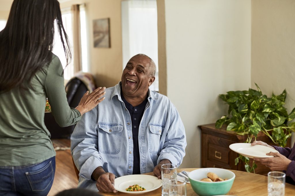 Un hombre se sienta a la mesa frente a un plato de verduras mientras sonríe a una mujer que pasa y le ha puesto una mano en el hombro derecho.