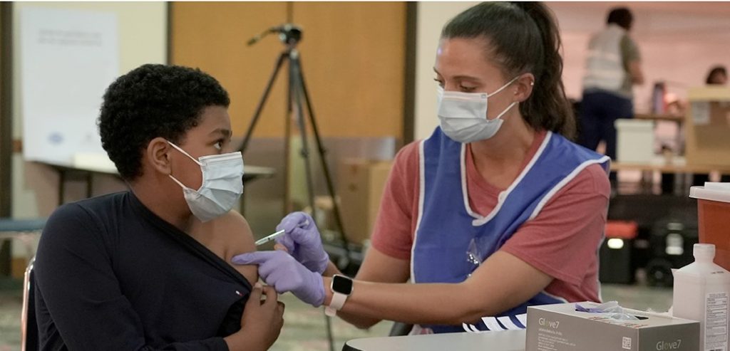 Una enfermera con máscara le aplica a un adolescente con máscara una inyección de COVID-19 en su hombro izquierdo.