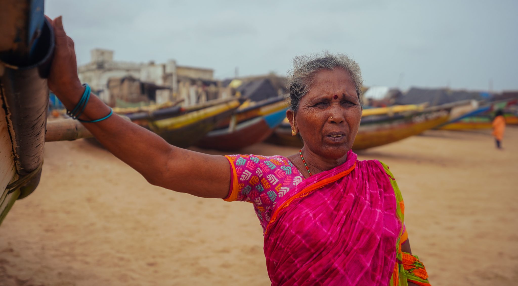 Una anciana viste un sari rosa brillante en la playa con barcos de pesca en el fondo