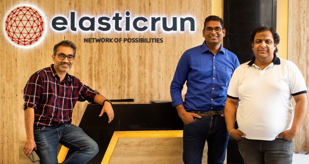 Tres hombres de pie frente a una pared con el logo de ElasticRun