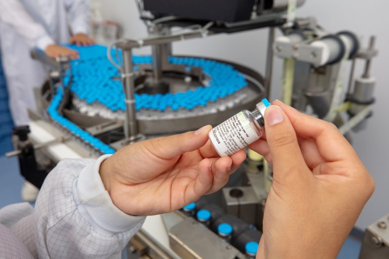 Un científico sostiene un frasco de medicamento con las dos manos, de pie en un laboratorio frente a un carrusel que contiene docenas de otros frascos.