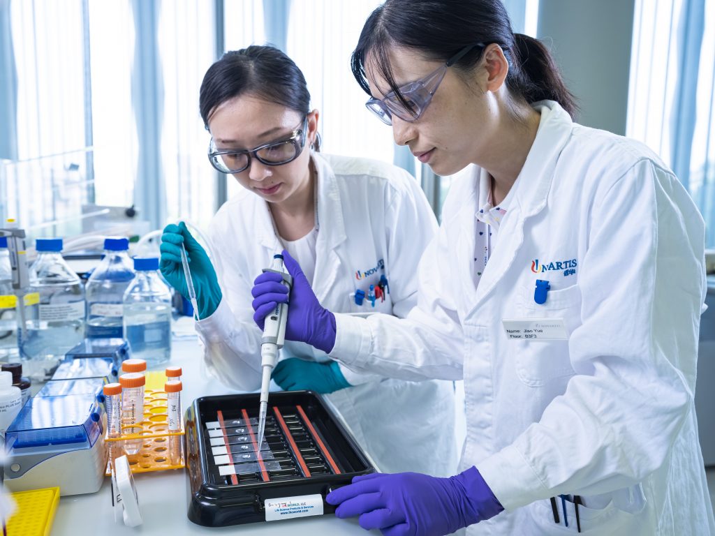 Dos científicas junto a una mesa de laboratorio; una de ellas aplica líquido a un portaobjetos de vidrio.