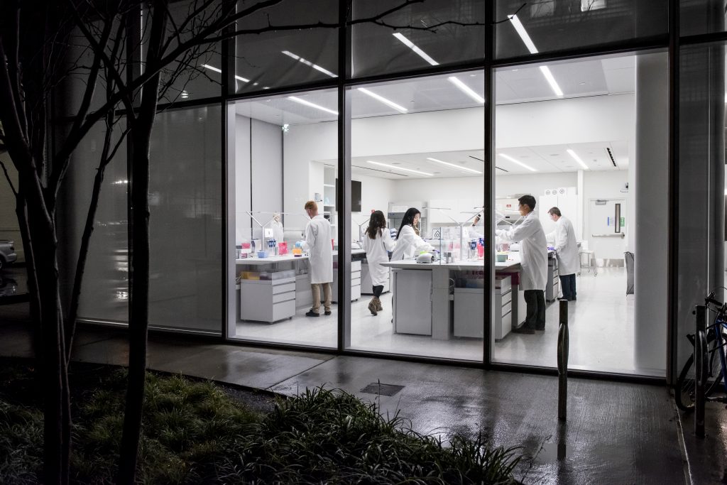 Cinco científicos de Novartis trabajan en bancos de laboratorio, como escena desde el exterior a través de una ventana.