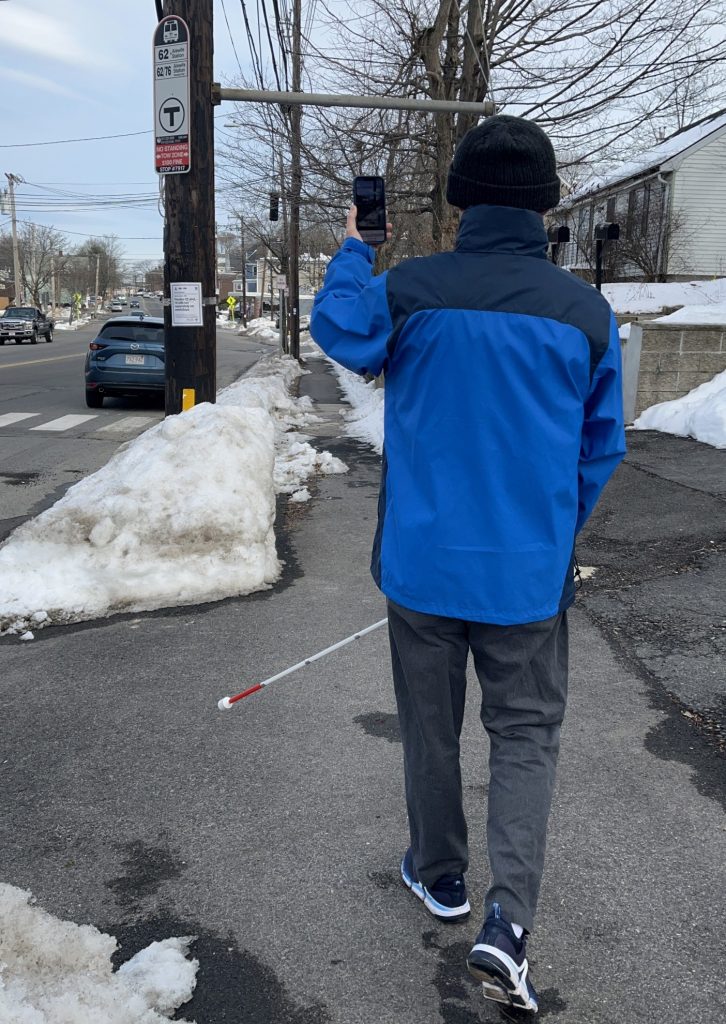 Hombre sostiene su bastón blanco en una mano y el teléfono en la otra con All Aboard activa para navegar hasta una parada de autobús