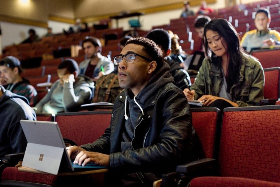 Primer plano de un estudiante universitario masculino sentado en la sala de conferencias de la universidad, mirando hacia adelante mientras toma notas en un Surface Pro. Otros estudiantes, hombres y mujeres, se sientan detrás de él y toman notas.