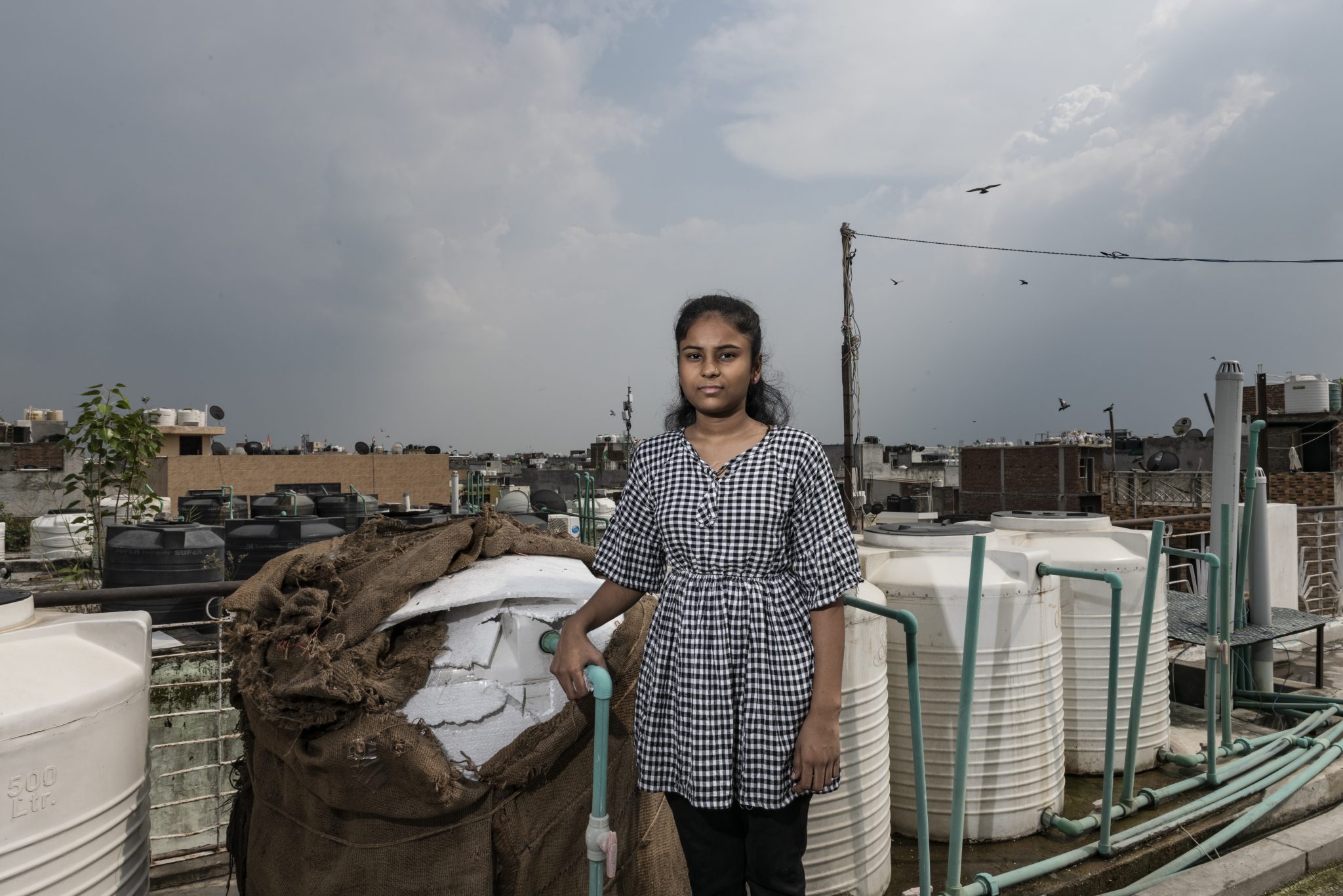 Una adolescente parada en la terraza de su casa junto a un tanque de agua cubierto con láminas de poliestireno y sacos de yute para aislarlo del calor